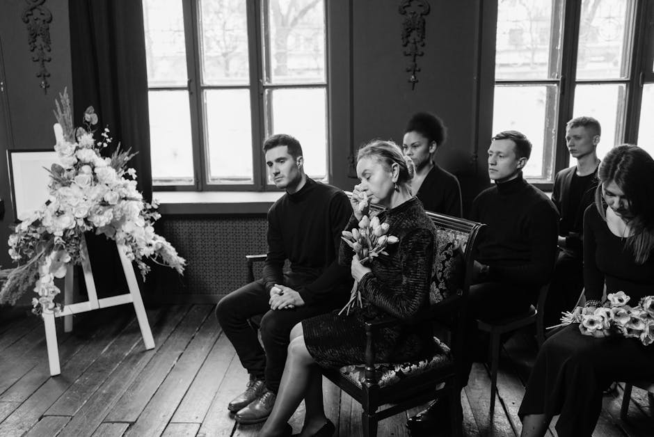 A black and white image showing six people attending a solemn funeral or memorial service indoors. They are seated in a row, with serious and contemplative expressions. The woman in the center holds a