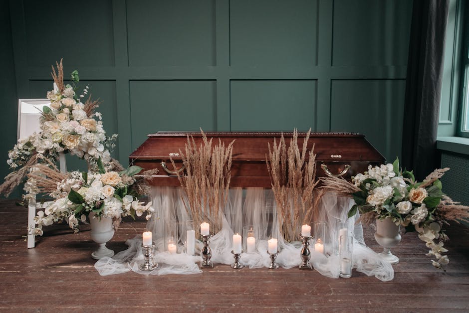 A floral arrangement for a funeral, featuring two large white vases filled with soft cream and white roses, hydrangeas, and greenery, placed symmetrically on either side of a polished wooden casket. T