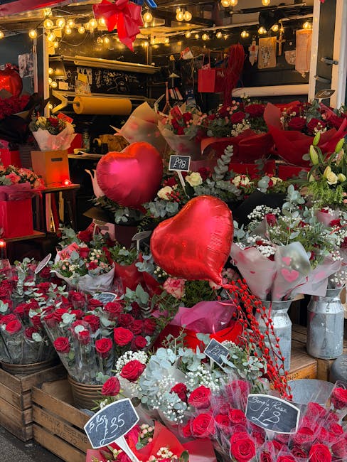 A vibrant display of floral arrangements at a flower shop, featuring predominantly red roses and white filler flowers, presented in glass jars, baskets, and wrapped bouquets. Brightly colored heart-sh