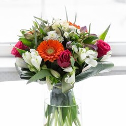 A lush tapestry of flowers comes to life in this Catford-inspired bouquet, masterfully arranged for balance and emotional resonance. At its heart, soft peach and fiery orange gerbera daisies radiate warmth-like a sunrise over Ladywell Fields-each petal unfurling with refined simplicity and energy. Deep fuchsia roses, densely petalled and stylishly nested, bring notes of romance and boldness, ensuring the bouquet captivates from every angle. Accenting the arrangement, ivory-white alstroemeria and miniature spray roses add texture and gentle brightness, interlaced with whispers of pastel pink blooms and fine, cloud-like white fillers that breathe light into the composition. A generous sweep of glossy, paddle-shaped aspidistra leaves wraps the stems, forming a vibrant collar that anchors the visual drama and gives the bouquet its signature round fullness. Varied greenery and slender sprigs fill the gaps, introducing movement and contrast. Arranged in a tall, clear glass vase that reveals every vibrant stem, the bouquet exudes freshness, as if gathered that morning from the local flower stalls near Catford Station. Soft, natural light illuminates the flowers, accentuating both subtle and dramatic tones. The effect is one of celebration and hope-ideal for a family gathering, a heartfelt thank-you, or simply brightening a home on a quiet afternoon in Catford, reflecting the careful attention only a true local florist brings.