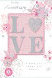 The anniversary card in this scene feels like a whisper of long-lasting romance, rendered in a palette of pastel pinks, silvery greys, and soft white that glows quietly against the surface beneath it. At its centre, the word "LOVE" stands out in raised, glitter-soft letters, each stroke of the typeface edged with a subtle shimmer. The letter "O" is transformed into a small, shining heart, textured with silver so it appears almost like a tiny polished charm set into the paper. Around this focal point, clusters of delicate, illustrated flowers unfold in blush, mauve, and gentle ivory tones, their lines fine and fluid, much like stems arranged thoughtfully in a vase on a Catford windowsill. Hints of butterflies and swirling flourishes thread through the design, adding an airy sense of movement and suggesting a breeze through a spring garden. The background, a soft pink wash, supports the artwork without ever overwhelming it, allowing the flowers and the glittered lettering to remain the true stars. In graceful, flowing script, the phrases "On Our Anniversary" and "I love you today and always" are set with plenty of breathing room, giving the eye-and the heart-space to rest. The composition feels intentionally balanced, elegant yet approachable, like a bespoke bouquet created for delivery around Catford and Bellingham. It conveys not just celebration, but the quiet security of shared years, making the card a tender companion to anniversary roses or a pastel arrangement chosen to honour a love that has grown richer over time.